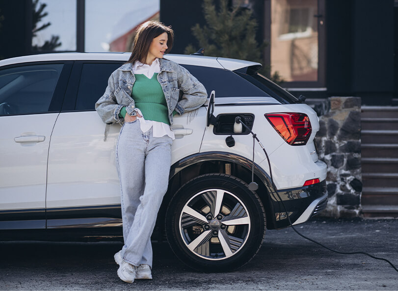 A young woman in front of a home, leaning against an electric vehicle while it charges.